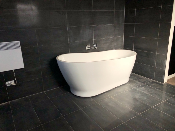A black tiled bathroom with a white free standing bath on the wall.